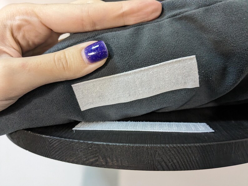 May include: Close-up of a black fabric cushion with two white hook and loop fasteners. The cushion is being held by a hand with purple nail polish.