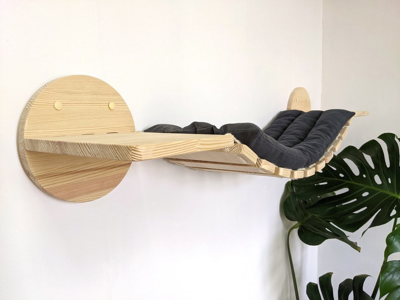 May include: A wooden cat bed with a black cushion, mounted on a wall. The bed is shaped like a hammock and has a circular platform on the left side.