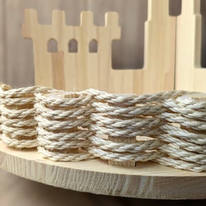 Op de afbeelding: Een houten plank in de vorm van een kasteel met een touwachtige rand rond de rand. De plank is gemaakt van licht hout en het touw is van natuurlijke beige kleur.