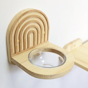 Op de afbeelding: Een houten voerbakhouder met een regenboogontwerp. De houder is aan een muur gemonteerd en heeft een glazen kom.