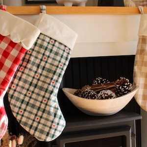 May include: Three fabric Christmas stockings hanging in a row. The first is red and white gingham with a scalloped white cuff. The second is green and white plaid with tiny red hearts and a straight white cuff. The third is beige and white gingham.