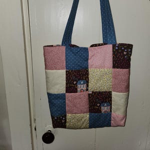 May include: A patchwork tote bag with a blue, pink, brown, and cream colour scheme. The bag has a quilted design and features small houses with white trim.