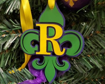 May include: A green and blue fleur-de-lis ornament with the letter "R" in yellow. The ornament is hanging on a green Christmas tree with purple ribbon and a purple ornament with a gold fleur-de-lis.