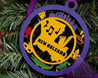 May include: A purple and gold Christmas ornament with a silhouette of the state of Louisiana, the words "New Orleans", and musical notes. The ornament is decorated with a fleur-de-lis, a crocodile, and other symbols of New Orleans.