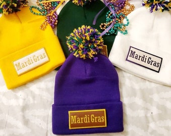 May include: Four Mardi Gras themed knit hats with pom-poms. Each hat has a different color: yellow, green, white, and purple. Each hat has a patch that says "Mardi Gras".