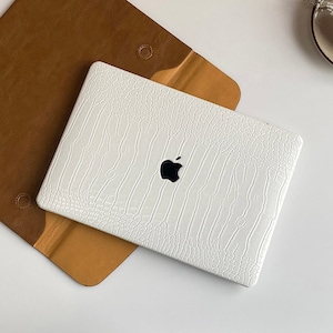 Puede incluir: Una funda para portátil blanca con un patrón de piel de cocodrilo y un logotipo de Apple. La funda está sobre una funda de cuero marrón.