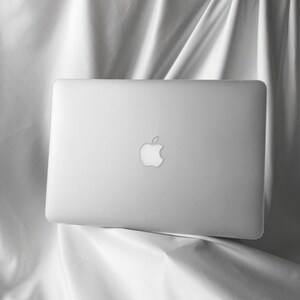 Könnte beinhalten: Ein silberner Laptop mit dem Apple-Logo auf dem Deckel. Der Laptop steht auf einer weißen Stoffoberfläche.
