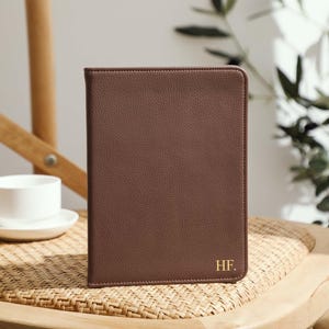 May include: A brown leather journal or notebook with the initials "HF." in gold lettering. The journal is closed and stands upright on a woven chair. A white teacup and saucer are in the background.