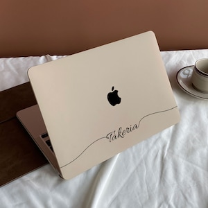 Puede incluir: Un ordenador portátil cerrado con una funda de cuero marrón. El portátil tiene una tapa blanca con un logotipo de Apple negro y la palabra "Takeria" escrita en cursiva.