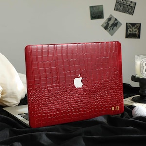 Può includere: Una custodia rossa per laptop con una texture a pelle di coccodrillo, con il logo Apple e le iniziali "R.B.". La custodia è su un laptop, con una candela e una pallina da golf nelle vicinanze. Lo sfondo include arte decorativa.