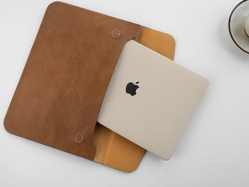 Puede incluir: Una funda de cuero marr&oacute;n con un interior de tela beige. La funda tiene una solapa que se cierra con un broche a presi&oacute;n. La funda est&aacute; dise&ntilde;ada para guardar una tableta o un port&aacute;til.