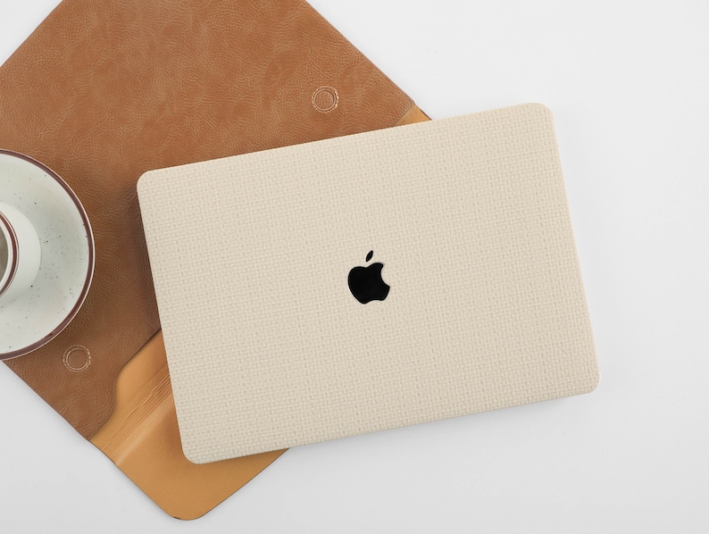 Puede incluir: Una funda para port&aacute;til beige con un logotipo de Apple negro. La funda tiene una superficie texturizada.