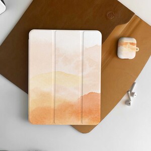 Puede incluir: Una funda blanca para tablet con un diseño de acuarela en tonos naranja y amarillo. La funda está hecha de tres piezas que encajan. La funda está sobre una superficie de cuero marrón.