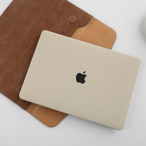 Puede incluir: Una funda para port&aacute;til beige con una solapa de cuero marr&oacute;n y un logotipo de Apple negro. La funda est&aacute; hecha de un tejido texturizado.