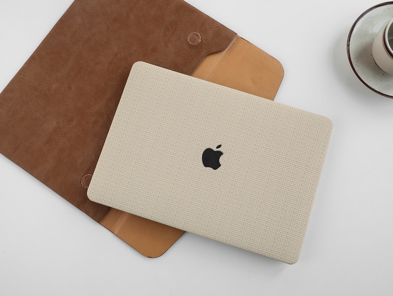Puede incluir: Una funda para port&aacute;til beige con una solapa de cuero marr&oacute;n y un logotipo de Apple negro. La funda est&aacute; hecha de un tejido texturizado.