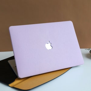 Puede incluir: Una funda para portátil de color lavanda con un logotipo de Apple plateado. La funda está cerrada y se encuentra sobre una superficie marrón.