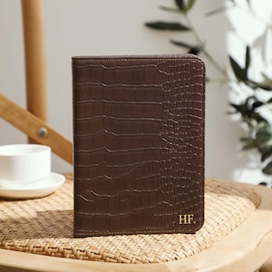 Op de afbeelding: Een bruin leren notitieboek met een krokodillenstructuur, gepersonaliseerd met de initialen "HF.". Het notitieboek is gesloten en staat rechtop op een geweven oppervlak. Een witte kop en schotel staan op de achtergrond.