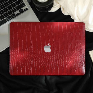 May include: A red laptop case with a crocodile skin texture. The case has a silver Apple logo in the center. The laptop is on a black surface next to a laptop keyboard and a book.