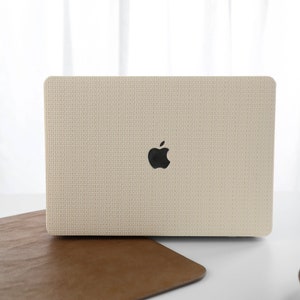 Puede incluir: Una funda para port&aacute;til beige con un acabado de tejido texturizado y un logotipo de Apple. La funda est&aacute; sobre una superficie de cuero marr&oacute;n.