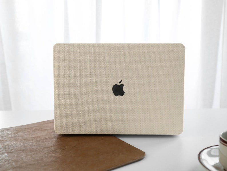 Puede incluir: Una funda para port&aacute;til beige con un acabado de tejido texturizado y un logotipo de Apple. La funda est&aacute; sobre una superficie de cuero marr&oacute;n.