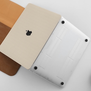 Puede incluir: Un ordenador port&aacute;til blanco con una funda de cuero beige. El port&aacute;til tiene un acabado plateado y un logotipo de Apple negro en la tapa. La funda tiene una superficie texturizada.