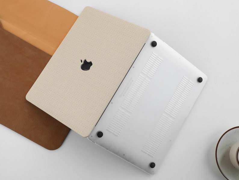 Puede incluir: Un ordenador port&aacute;til blanco con una funda de cuero beige. El port&aacute;til tiene un acabado plateado y un logotipo de Apple negro en la tapa. La funda tiene una superficie texturizada.
