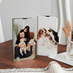 May include: Two phone cases with different designs. One case features a photo of a family with a baby, while the other case features a photo of a dog and a cat. Both cases are white with a textured surface.