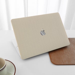 Puede incluir: Una funda para port&aacute;til beige con un acabado de tejido texturizado y un logotipo de Apple. La funda est&aacute; abierta y el port&aacute;til es visible en el interior.