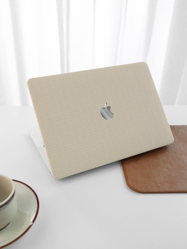 Puede incluir: Una funda para port&aacute;til beige con un acabado de tejido texturizado y un logotipo de Apple. La funda est&aacute; abierta y el port&aacute;til es visible en el interior.