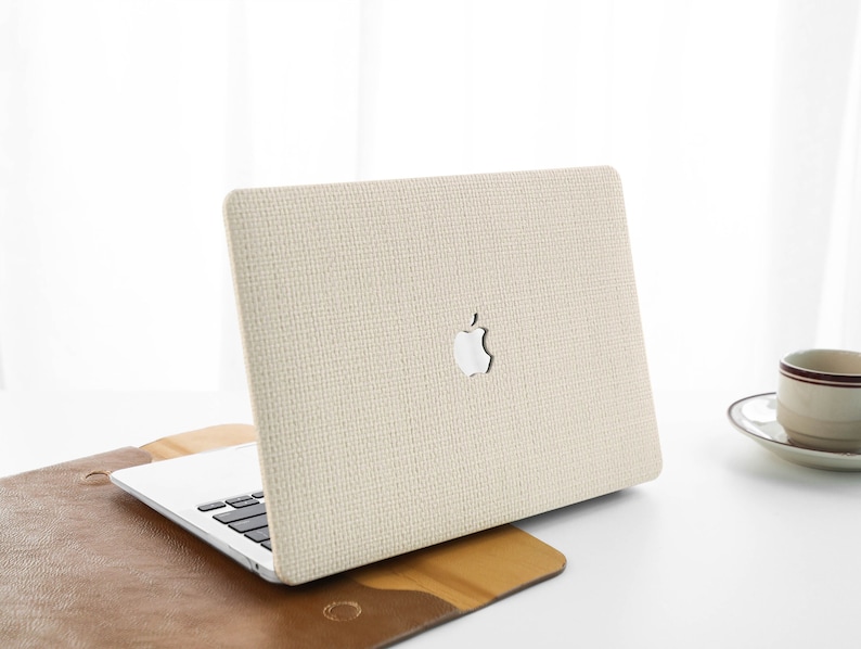 Puede incluir: Un ordenador port&aacute;til beige con una cubierta de tela texturizada y el logotipo de Apple en la tapa. El port&aacute;til est&aacute; sobre una superficie de cuero marr&oacute;n.