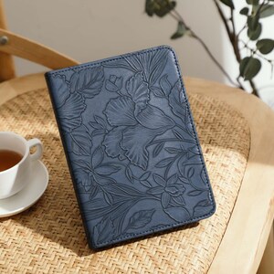 May include: A navy blue leather journal with a floral embossed design rests on a woven chair. A white teacup and saucer with tea are next to the journal. The journal's cover features a detailed pattern of flowers and leaves.