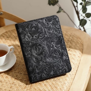 May include: Black leather journal with an embossed floral pattern. The journal is rectangular and sits on a woven chair. A white teacup and saucer with tea are beside the journal.