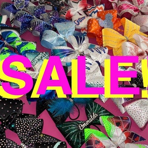 May include: A variety of cheer bows with rhinestones and different designs, including a green bow with a white and green design, a blue bow with a white and blue design, a red bow with a white and red design, a yellow bow with a white and yellow design, and a black bow with a white and black design. The bows are arranged in a random pattern on a pink surface. The word "SALE!" is written in large, pink letters across the center of the image.