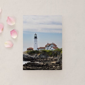 May include: A white lighthouse with a red roof stands on a rocky cliff overlooking the ocean. The sky is cloudy and blue.