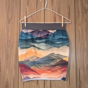 May include: A short skirt with a colorful mountain landscape print. The skirt has a gray waistband and a white hem.