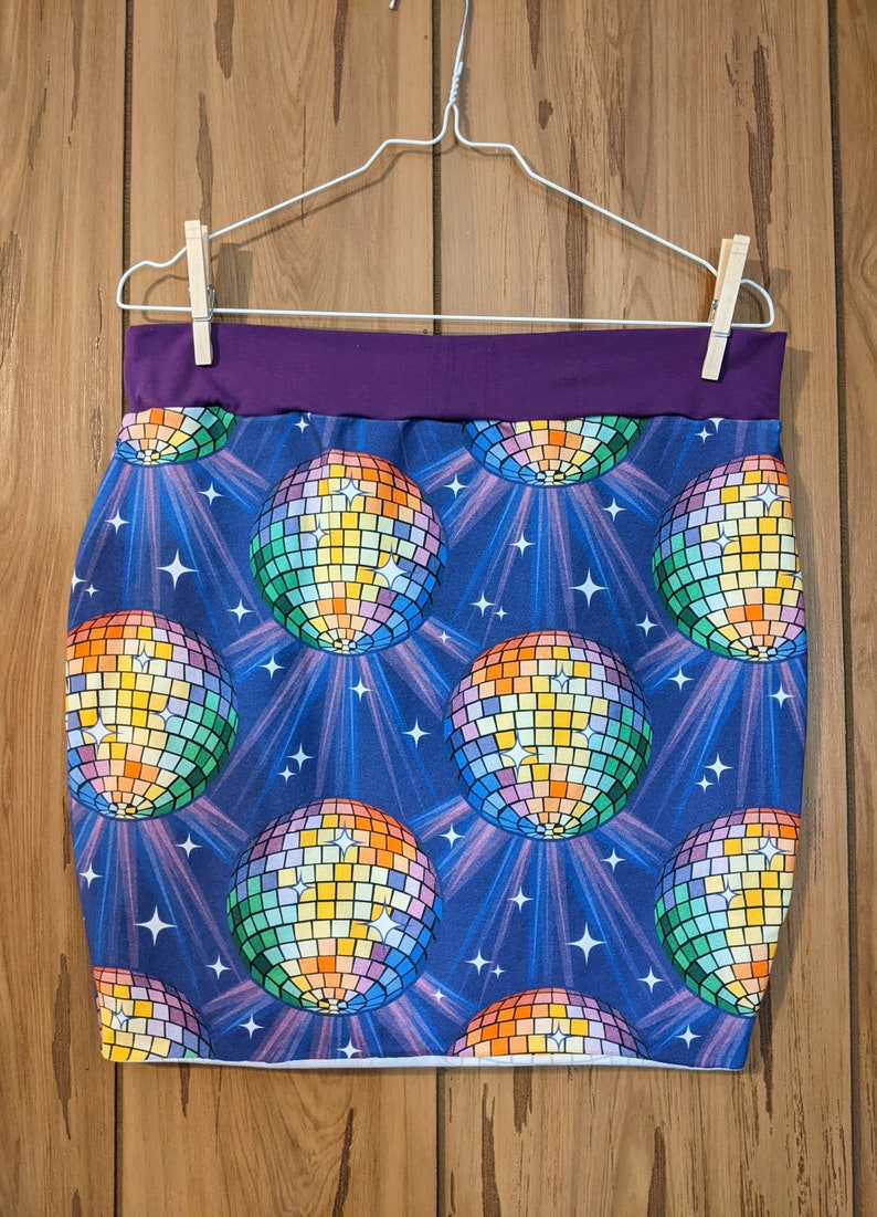 May include: A purple waistband mini skirt with a blue background and a repeating pattern of rainbow disco balls. The disco balls are surrounded by white stars and white rays of light.