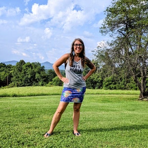 Puede incluir: Una mujer con una camiseta gris sin mangas con el texto "Let's Dance" y una falda azul con estampado de bola de discoteca se encuentra en un campo de hierba. Est&aacute; sonriendo y mirando a la c&aacute;mara.