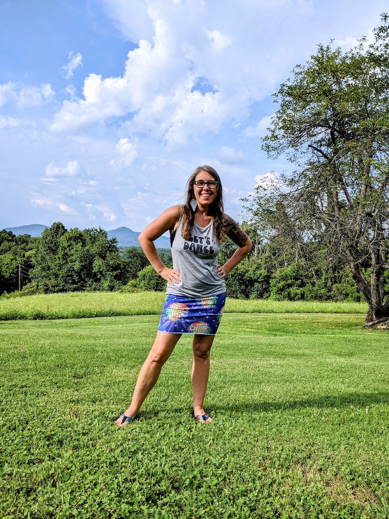 Puede incluir: Una mujer con una camiseta gris sin mangas con el texto "Let's Dance" y una falda azul con estampado de bola de discoteca se encuentra en un campo de hierba. Est&aacute; sonriendo y mirando a la c&aacute;mara.