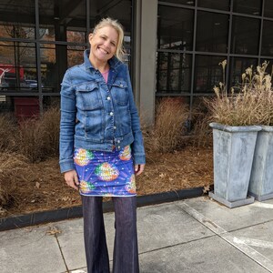 Puede incluir: Una mujer con una chaqueta vaquera azul, una falda con estampado de bola de discoteca de colores y unos pantalones vaqueros acampanados de color azul oscuro. Est&aacute; de pie frente a un edificio con grandes ventanales.