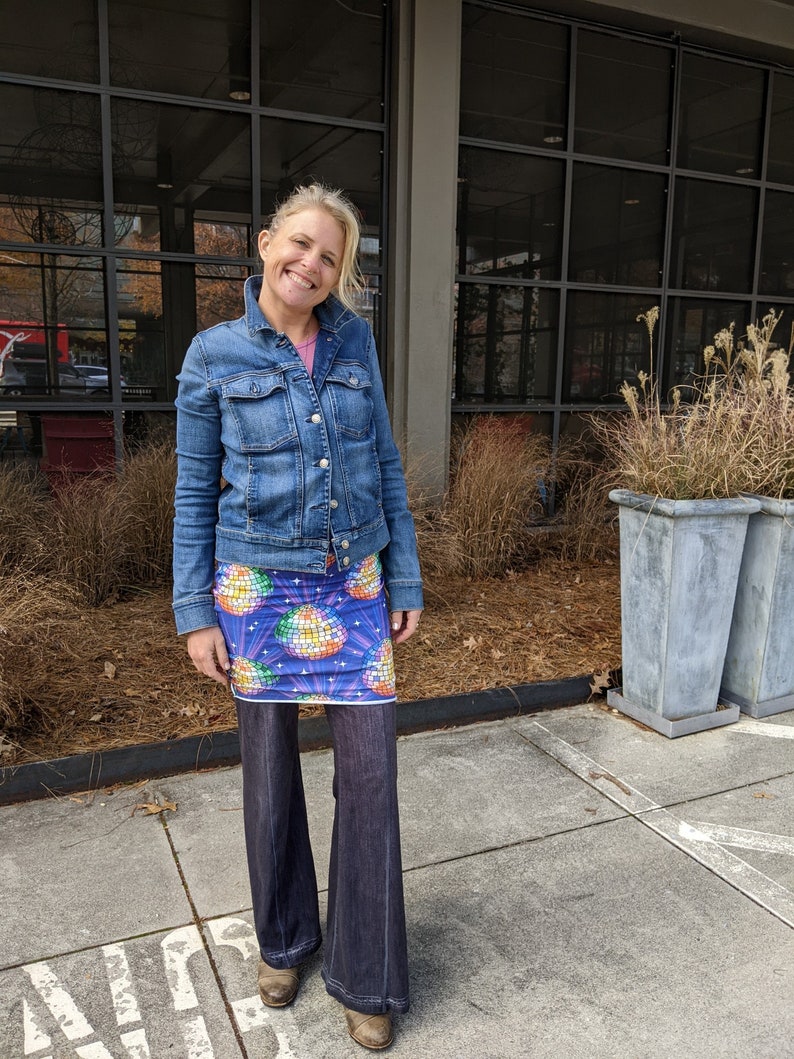 Puede incluir: Una mujer con una chaqueta vaquera azul, una falda con estampado de bola de discoteca de colores y unos pantalones vaqueros acampanados de color azul oscuro. Est&aacute; de pie frente a un edificio con grandes ventanales.