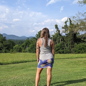 Puede incluir: Una mujer con un top gris sin mangas y una falda colorida con estampado de pi&ntilde;a se encuentra de pie en un campo de hierba, mirando hacia una cordillera en la distancia.