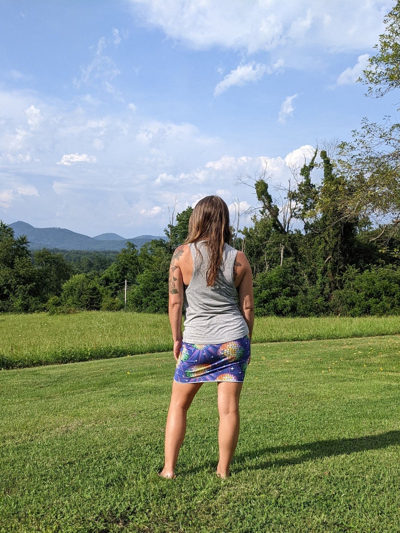 Puede incluir: Una mujer con un top gris sin mangas y una falda colorida con estampado de pi&ntilde;a se encuentra de pie en un campo de hierba, mirando hacia una cordillera en la distancia.