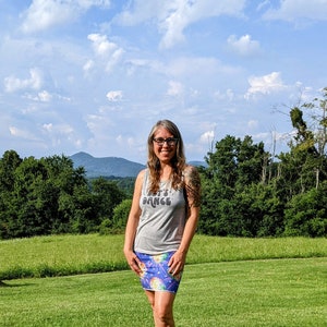 Puede incluir: Una mujer lleva una camiseta sin mangas gris con el texto "Let's Dance" y una falda colorida con un estampado de bola de discoteca. Est&aacute; de pie en un campo de hierba con una cordillera de monta&ntilde;as en el fondo.