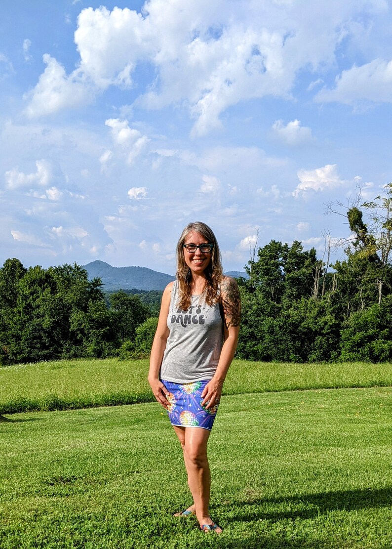 Puede incluir: Una mujer lleva una camiseta sin mangas gris con el texto "Let's Dance" y una falda colorida con un estampado de bola de discoteca. Est&aacute; de pie en un campo de hierba con una cordillera de monta&ntilde;as en el fondo.