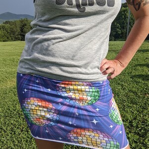 Puede incluir: Una falda corta con fondo azul y un patr&oacute;n de bola de discoteca. La falda tiene un ribete blanco y est&aacute; hecha de una tela el&aacute;stica. El texto "Let's Dance" es visible en una camiseta sin mangas gris.