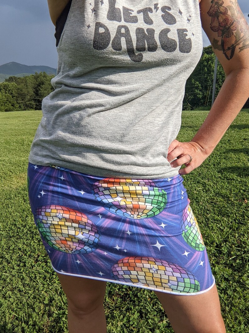 Puede incluir: Una falda corta con fondo azul y un patr&oacute;n de bola de discoteca. La falda tiene un ribete blanco y est&aacute; hecha de una tela el&aacute;stica. El texto "Let's Dance" es visible en una camiseta sin mangas gris.