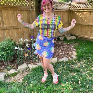 Puede incluir: Una mujer con un top corto de tie-dye arco&iacute;ris con el texto "The Sloth" y una falda a juego de discoteca arco&iacute;ris y morada. Lleva sandalias blancas con suelas rojas, tiene el pelo rosa y gafas de sol.