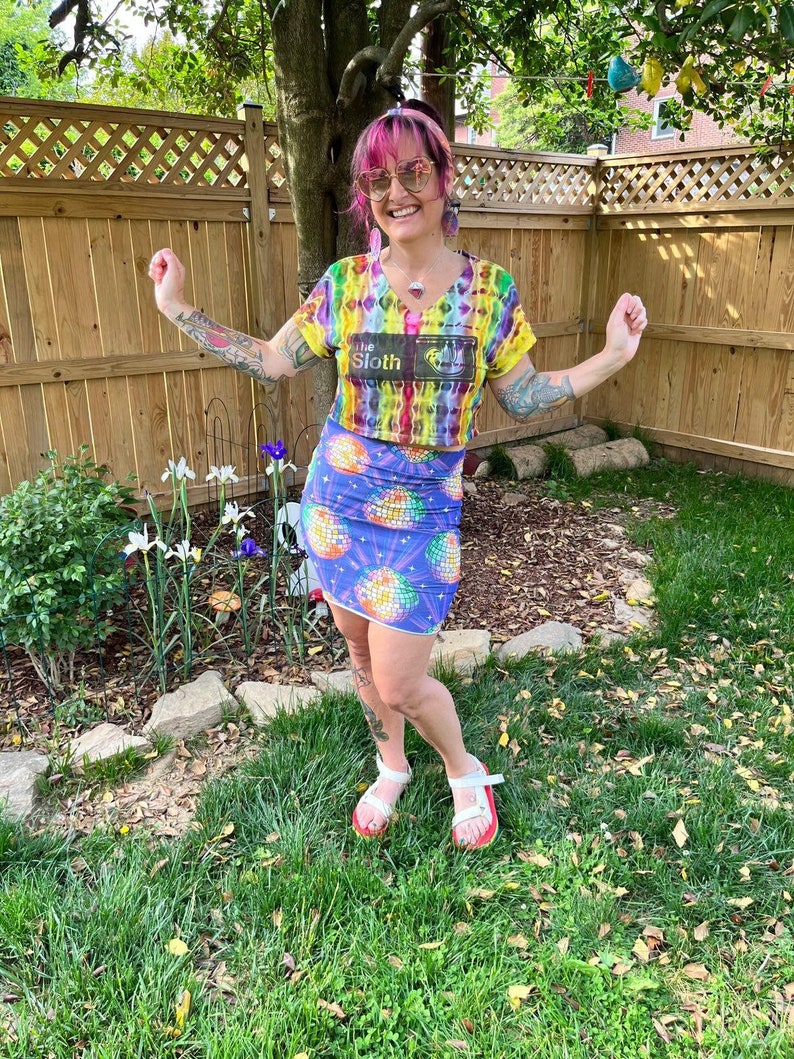 Puede incluir: Una mujer con un top corto de tie-dye arco&iacute;ris con el texto "The Sloth" y una falda a juego de discoteca arco&iacute;ris y morada. Lleva sandalias blancas con suelas rojas, tiene el pelo rosa y gafas de sol.