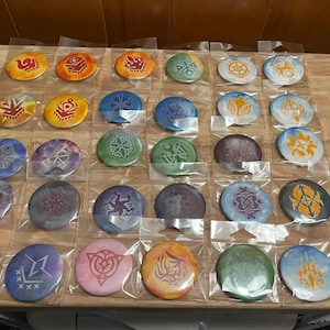 May include: A collection of 25 round pinback buttons with various colorful designs. The buttons feature different symbols and patterns, including geometric shapes, stars, and abstract designs.