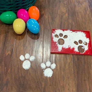 May include: A red plastic paw print stencil with white flour dusted on top. Four paw prints are visible on the wooden floor, made with the stencil.